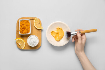 Female hand with brush, bowl with turmeric mask, powder and lemon on grey background