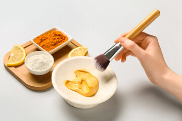 Woman with makeup brush taking turmeric mask from bowl on grey background