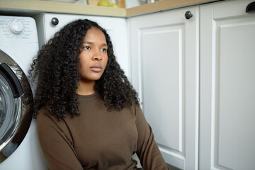 Portrait of young beautiful sad thoughtful African American female, sitting at kitchen, on floor, leaning against white furniture, having lovely fuzzy hair. Emotions and feelings, face expressions