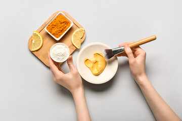 Woman with makeup brush making turmeric mask in bowl on grey background