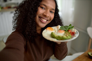 Happy smiling woman of black ethnicity with curls showing her colorful delicious and healthy breakfast with eggs, avocado and greens at camera, doing selfie for her social network page