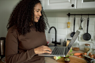 Pretty happy dark-skinned female in brown clothes holding laptop, videochatting with her friend while cooking breakfast in late morning of lazy weekend, standing next to table top with vegetables