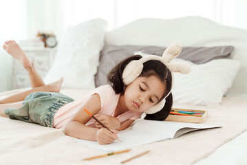 Cute little girl having fun in cute kindergarten age. Use colored pencils to draw a picture on the paper. Happily put the rabbit headphones on the bed. Concept family and happy learning