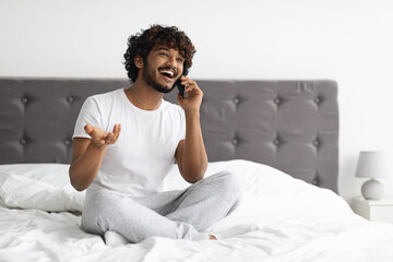 Positive hindu guy sitting on bed, talking on phone