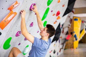 Fototapeta premium European young man grabbing ledges of artificial climbing wall in bouldering centre.