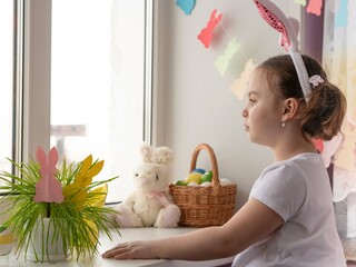 A little girl with rabbit ears in a white sweater stands hopefully looking out the window with her...