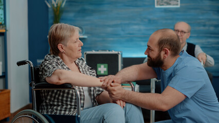 Man nurse holding hands and comforting pensioner with disability in nursing home. Medical assistant having conversation with elder disabled woman about rehabilitation and healthcare.