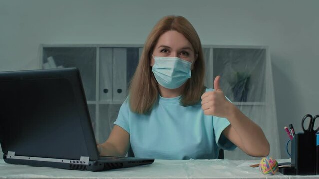Middle aged businesswoman in medical mask sitting working typing email on laptop using new computer app showing thumbs up gesture approval sign affirmation happy with modern technology smiling