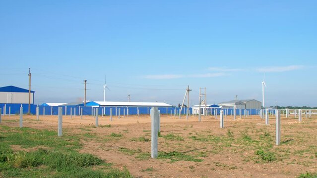 Vertical bars for sun panels installation at solar station in countryside. Powerful wind turbines generate alternative electric power against blue sky