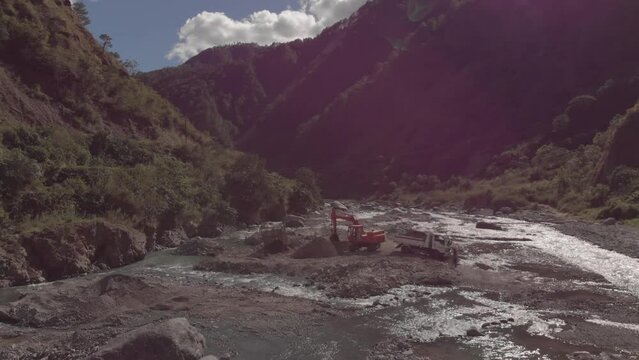 excavator digging rocks out of river bed in mountainous region in Benguet Kabayan Philippines wide angle aerial approaching raw cement material mining flowing water reflecting light bright sunny day