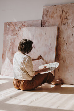 Elegant woman artist sitting on the floor in studio with sunlight shadows and painting abstract artwork