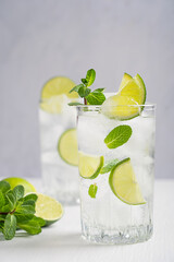 Homemade cold refreshing lemonade drink or cocktail made of sparkling water, lime slices and fresh green mint leaves served in tall drinking glass with ice cubes on white wooden table at summer