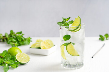 Cold homemade lemonade or cocktail made of tonic, lime slices and fresh green mint leaves served in tall drinking glass with ice cubes on white wooden table with ingredients for refreshment at summer