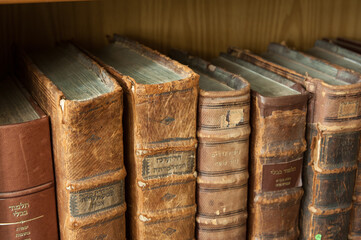Dusty, worn and faded leather-bound Jewish holy books stacked upright on a library shelf.