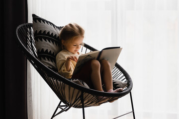 little girl sitting in a chair by the window reading a book