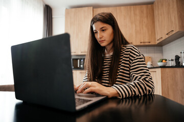 Fototapeta premium Young female freelancer dressed in home clothes working on laptop sitting at the table in the kitchen. Stylish girl on workplace at the desk. Distance learning online education