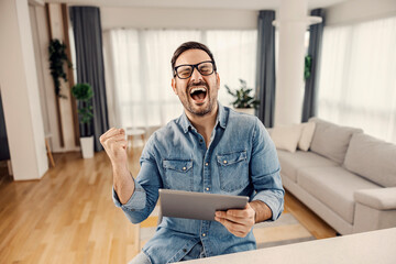An excited man holding a tablet and celebrating success or good news at his cozy home.