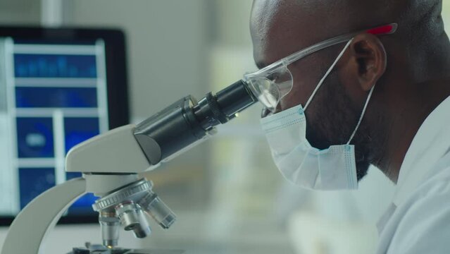 African American scientist in protective face mask and glasses looking through microscope while doing research in laboratory