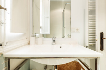 White porcelain sink on stainless steel metal structure with frameless square mirror and white heated towel rail behind the door