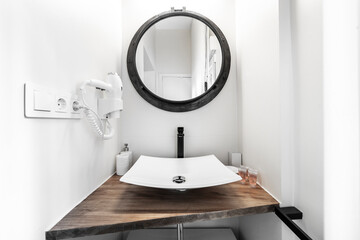 Small bathroom with wooden countertop, porcelain sink without insert, round mirror with black frame and white hair dryer on the wall