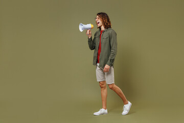 Fototapeta na wymiar Full size body length side profile view young brunet curly man 20s wear khaki shirt hold scream in megaphone announces discounts sale Hurry up isolated on plain olive green background studio portrait