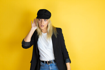 Beautiful woman wearing casual white t-shirt and a cap over isolated yellow background surprised with hand over ear listening an hearing to rumor or gossip