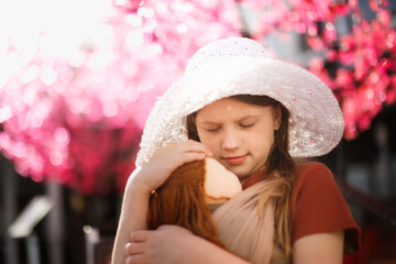 Cute long-haired european kid girl with doll in sling, spring photography. Girl with doll on background of flowering trees. conscious natural parenting, doll play