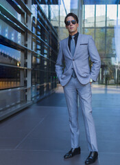 full length photo of an elegant man in sunglasses and formal business wear with his hands in the pockets looking serious at the camera while is standing on an entry of a offices building