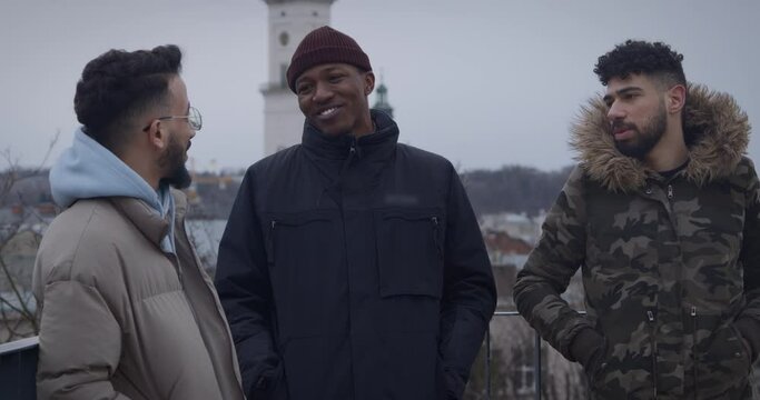 Cheerful multiracial male friends meet in city center to talk on street. Arabian with African American and Moroccan man against church in old town closeup