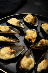 baked mussels in shells with creamy sauce poured with oil on a black plate on a dark background