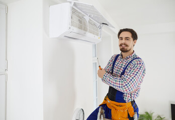 Happy repairman or technician repairing modern AC unit. Handsome young man standing on ladder with arms crossed after fixing problems with air conditioner, holding screwdriver and looking at camera