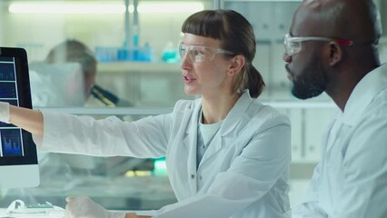 Caucasian female scientist and her African American male colleague in protective glasses and medical gloves discussing micrograph and infographics on computer while cooperating in laboratory - Powered by Adobe