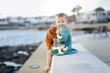 cute european baby toddler in a green dress with a doll sits on a stone bench by the sea, gray background. Girl playing with a doll in summer alone
