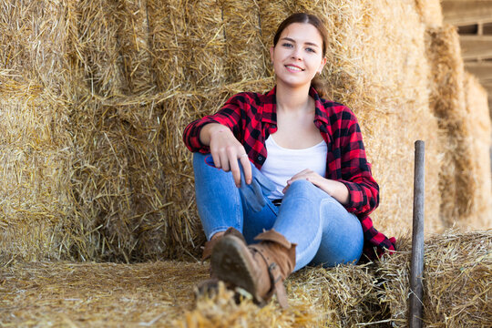 Young attractive female farmer resting on hayloft after work at farm, sitting on straw stack and smiling