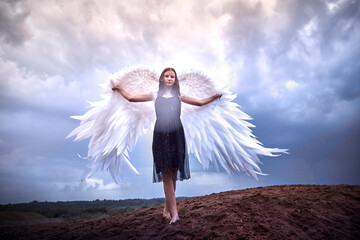 Young slim girl in black dress with with white wings dances on sand dunes against a dramatic sky...