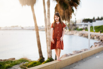 Cute long-haired European kid girl in a dress on the sea coast. A teenager child walks along a...