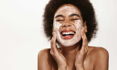 Spa and cosmetology. Young african american woman smiling happy laughing while washing face, cleansing skin, apply moisturizing cream, face lotion or toner, pink background
