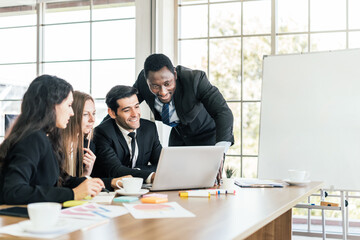 African businessmen standing in the back of Caucasian colleagues with smiles looking on laptop at an internal business group meeting with an empty whiteboard. Diversity of business people cooperation.