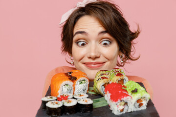 Close up shocked surprised amazed impressed satisfied young woman hold in hand look at makizushi sushi roll served on black plate traditional japanese food isolated on plain pastel pink background