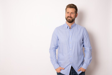 Fototapeta premium Portrait of charming successful young man in blue shirt, smiling broadly with self-assured expression while holding hands in pockets over white background. Business concept.