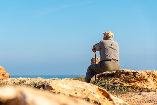 Pensive old man with beret sitting and with walking stick on the shore looking at the sea. Traditional lord of Spanish culture