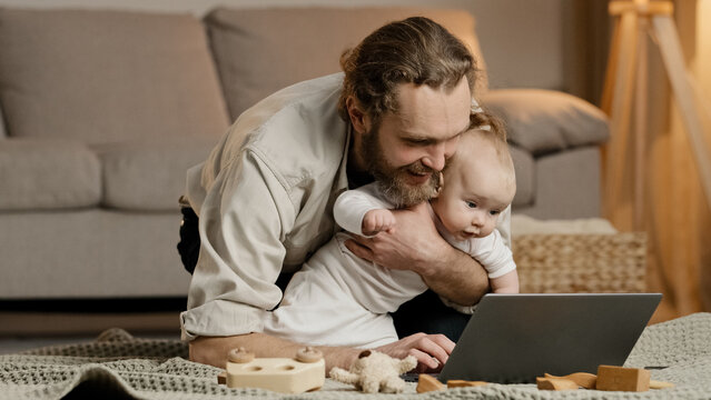 Single Father Bearded Man Dad With Little Girl Daughter Son Child Baby Kid Infant Newborn Sits At Home On Floor Looking At Laptop Video Call Browsing Online Uses Computer App Buys Baby Products Online