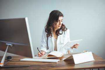 Doctor working at office desk. Female doctor sitting at the table writing in documents and working on laptop computer at office. Female doctor in a modern office clinic / hospital