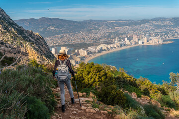 Fototapeta na wymiar A young woman descending from the top of the Penon de Ifach Natural Park with the city of Calpe in the background, Valencia. Spain. Mediterranean sea. View of La Fossa beach