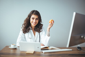 Portrait of beautiful smiling nutritionist looking at camera and showing healthy vegetables in the consultation. Portrait of young smiling female nutritionist holding apple in the consultation room