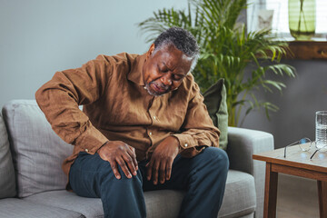 Photo of mature, elderly man sitting on a sofa in the living room at home and touching his knee by...