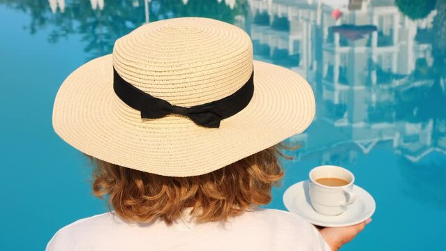 female 50 years old in a straw hat wearing a white dress relaxing by the pool with a cup of coffee, vacation concept, good morning. woman sitting by the pool.