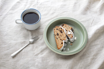 グレーのテーブルクロスに置かれた丸いカーキ色のお皿の上のシュトーレンとフォークとコーヒー