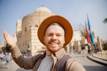 Young archaeologist man in hat takes selfie photo against background of ancient buildings. Concept archeological sit old city
