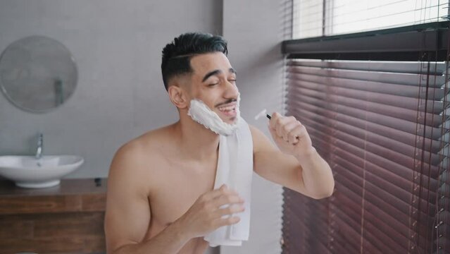 Happy singing dancing funny handsome naked arab man guy with white soap suds on beard active male in bath sings song into razor having fun shaving in bathroom at home dance music fun hygiene ritual
